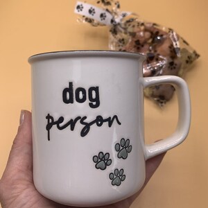 May include: A white wooden crate with a dog-shaped cutout and the word "SULLY" on the front. The crate contains a white mug with the text "dog person" and a bag of dog treats. The crate is on a light brown surface.