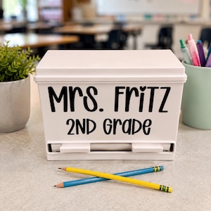 Op de afbeelding: Een witte rechthoekige plastic opbergdoos met deksel, voorzien van de zwarte tekst "Mrs. Fritz 2nd Grade" op de voorzijde. De doos heeft een horizontale gleuf aan de onderkant en er liggen twee potloden gekruist voor de doos.