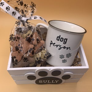 May include: White gift basket with a white mug that says "dog person" and a bag of dog treats. The basket is decorated with a paw print and the name "SULLY".