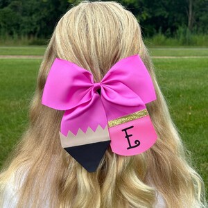 May include: A pink bow with a black and beige pencil design and a gold glitter stripe. The pencil design has a pink eraser with the letter "E" written on it.