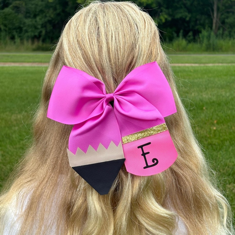 May include: A pink bow with a black and beige pencil design and a gold glitter stripe. The pencil design has a pink eraser with the letter "E" written on it.