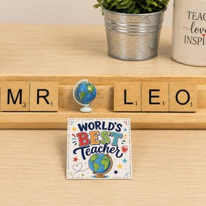 May include: A wooden tile rack displaying the letters M, R, L, E, O with a miniature globe between the words. A square card in front features the text WORLD'S BEST TEACHER with a globe illustration, colorful stars, and a small red heart.