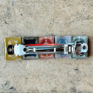 May include: A silver hair clip with a decorative design. The clip is made up of five small square tiles with different patterns. The tiles are in shades of yellow, red, blue, and black.