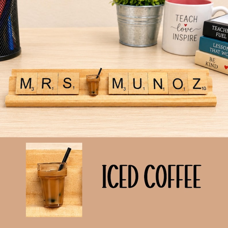 Op de afbeelding: Een houten tegelhouder met de letters M, R, S, M, U, N, O, Z en een miniatuur ijskoffiebeker met een zwart rietje die de letter A vervangt. De tegels zijn beige met zwarte letters en kleine numerieke waarden in de hoeken.