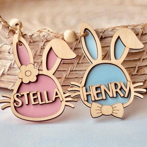 May include: Two wooden bunny-shaped Easter decorations with the names Stella and Henry. The bunny with the name Stella is pink and the bunny with the name Henry is blue.