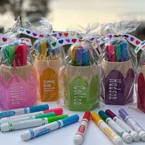 May include: Six colorful pencil-shaped containers with personalized names, Frankie, Olivia, Kali, Colton, Hudson, and Ezra, filled with colorful markers. The containers are arranged in a row on a white surface with additional markers scattered around them.