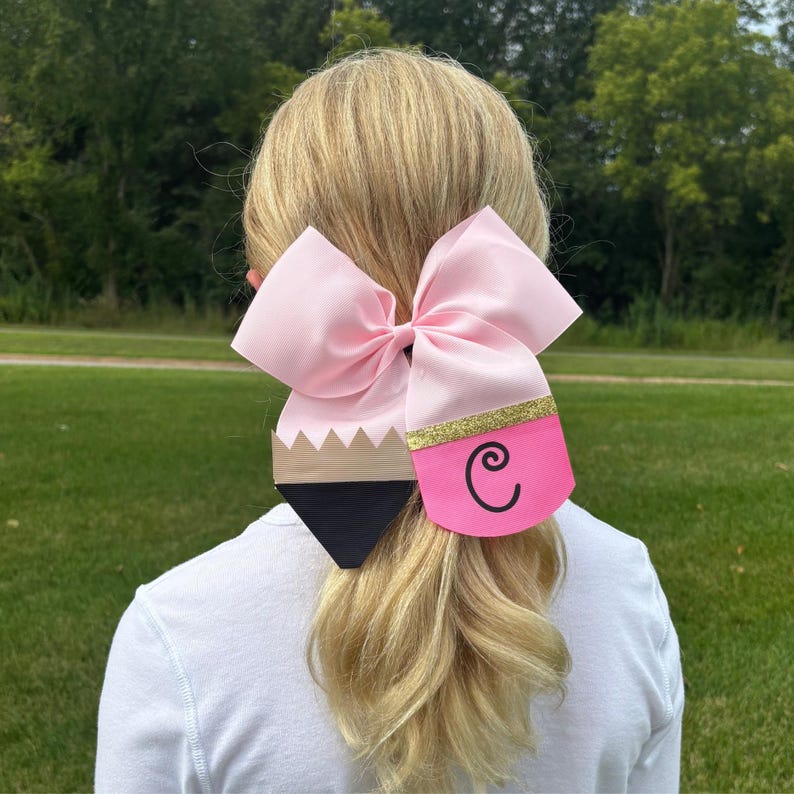 May include: A pink bow with a pencil-shaped embellishment. The pencil is black and brown with a gold glitter accent and the letter "C" on the pink eraser.