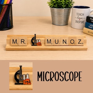 Op de afbeelding: Een houten tegelhouder met de naam MR. MUNOZ, met een metalen emaille speld van een zwarte microscoop en oranje met vloeistof gevulde bekers tussen de letters geplaatst. De tegels zijn voorzien van zwarte letters op een lichte houten achtergrond.