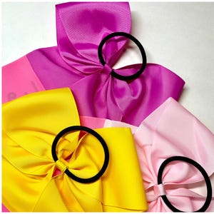 May include: Three hair bows in pink, yellow, and light pink with black elastic bands. The bows are made of grosgrain ribbon and are arranged in a stack.