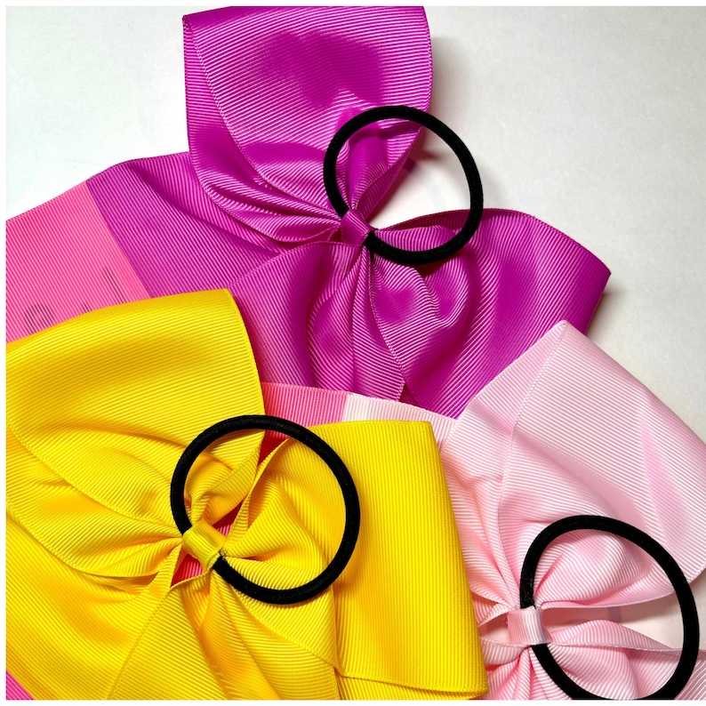 May include: Three hair bows in pink, yellow, and light pink with black elastic bands. The bows are made of grosgrain ribbon and are arranged in a stack.