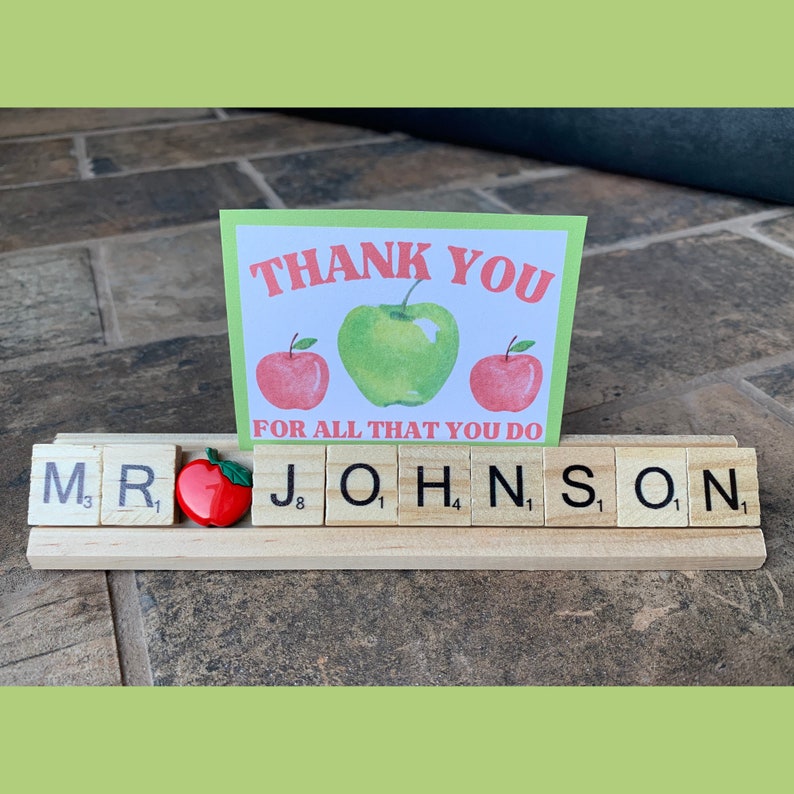 A wooden letter tile holder with the name 'MR JOHNSON' spelled out in black letter tiles. There is a red apple between the 'R' and 'J' tiles. A green card with red apples and a green apple with the text 'THANK YOU FOR ALL THAT YOU DO' is behind the letter tiles.