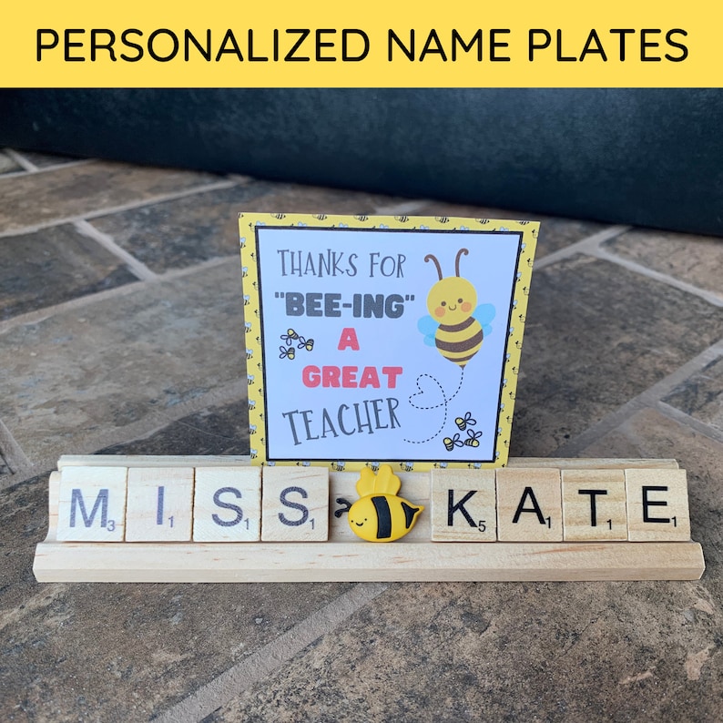 A wooden name plate with the word 'MISS' spelled out in white letters on individual wooden tiles. The tiles are arranged on a wooden base. A small yellow and black bee figurine is placed between the 'S' and 'K' tiles. A yellow card with a bee illustration and the text 'Thanks for 'Bee-ing' a Great Teacher' is propped up behind the name plate.