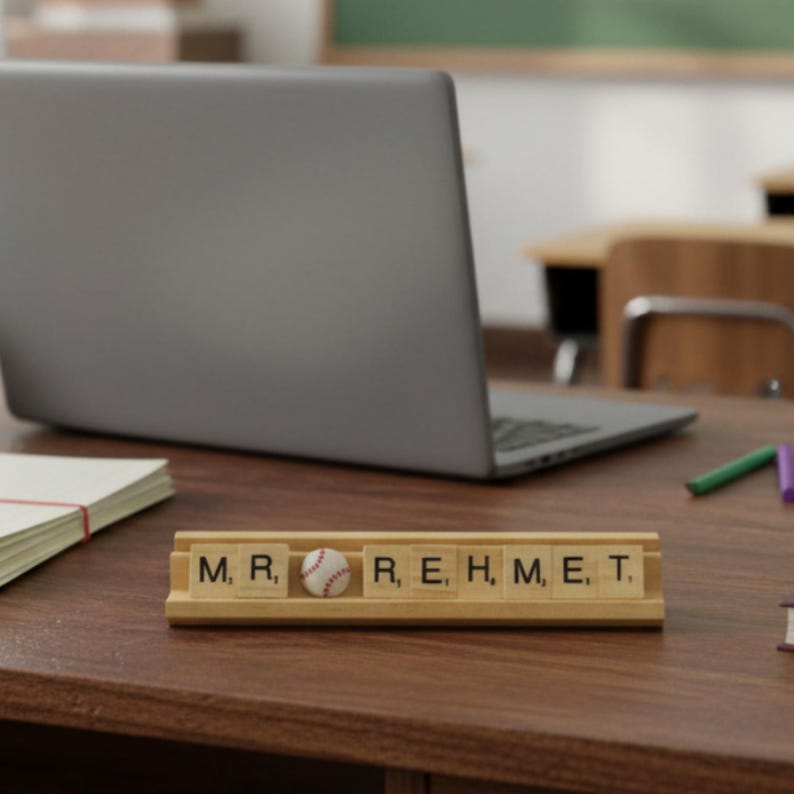 Op de afbeelding: Een houten tegelhouder toont de letters M, R, R, E, H, M, E, T met een kleine honkbal die de letter A vervangt. De tegels zijn op een rij geplaatst op een bureau voor een gesloten laptop, waardoor de naam MR. REHMET wordt gevormd.
