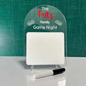 Könnte beinhalten: Klarer Familien-Spielabend-Schild aus Acryl mit dem Text "The Fritz family Game Night" in Rot und Weiß. Enthält eine weiße, trocken abwischbare Tafel und einen schwarz-weißen Marker. Würfelgrafiken sind ebenfalls enthalten.