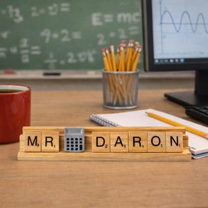 Op de afbeelding: Een houten bureau-naambordje met de tekst MR. DARON en een klein grijs rekenmachine-icoontje tussen de naamdelen. Het bordje staat op een bureau naast een rode koffiemok, een pennenhouder met gele potloden, een notitieblok en een computermonitor.