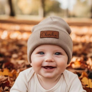 May include: A beige beanie featuring a brown leather patch with the name "Charlie" in cursive. The beanie is worn by a smiling baby, surrounded by autumn leaves. The baby is wearing a cream-coloured onesie.