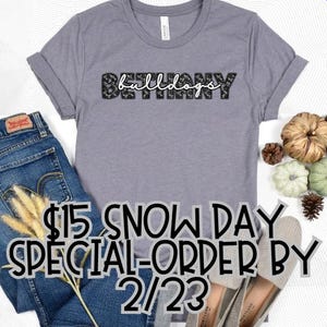 May include: A gray t-shirt with the text "Bethany Bulldogs" in black with a leopard print design. The shirt is on a white surface with a pair of jeans and some decorative gourds.