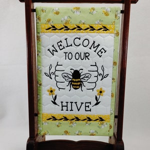 May include: A rectangular quilted fabric sign hanging on a dark brown wooden stand. The sign features a light green background with a bee pattern, a white center panel with a honeycomb stitch, and the black embroidered text "WELCOME TO OUR HIVE".