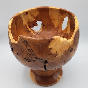 May include: A handcrafted wooden bowl with a pedestal base. The bowl features a natural wood grain pattern with light and dark brown hues. The bowl has several openings and a unique, organic shape.