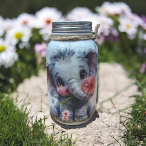 May include: A glass jar with a silver lid and a decorative rope around the neck. The jar is adorned with a charming illustration of a baby elephant holding a flower. The elephant is light blue with pink accents, and the jar is surrounded by flowers.