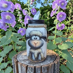 May include: A mason jar tissue holder featuring a whimsical sheep design. The jar is painted white with a black lid and a twine accent. The sheep has a fluffy white face, black ears, and a cute expression. The tissue holder is sitting on a wooden stump.