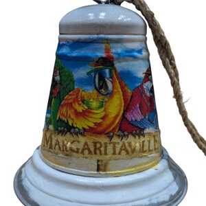 May include: White bell with a distressed finish, featuring a colorful parrot design and the word "Margaritaville." The bell has a rope for hanging.