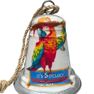 May include: A decorative bell with a parrot design. The bell is white with a colorful parrot illustration in red, yellow, blue, and green. The text "IT'S 5 O'CLOCK SOMEWHERE" is displayed on a blue banner. A rope is attached for hanging.