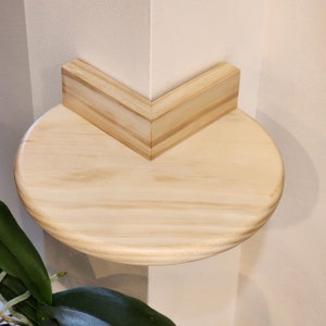 May include: A round wooden shelf with a natural finish, mounted in a corner of a wall. The shelf is made of light wood and has a rounded edge.