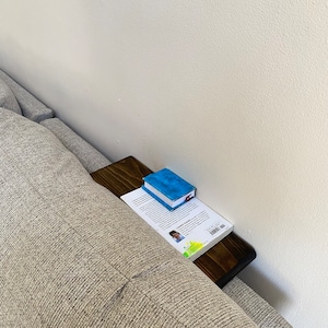 Puede incluir: Una mesa auxiliar de madera unida a un sofá gris con un libro y un pequeño libro azul en la parte superior.