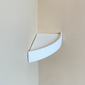 May include: A white corner shelf with a curved front edge. The shelf is mounted in the corner of two walls painted in a neutral beige color. The shelf is designed to fit snugly into the corner.