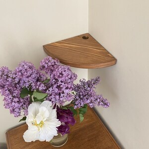 Puede incluir: Una estantería de esquina de madera con un tinte marrón claro. La estantería está montada en una pared blanca y tiene un ramo de flores moradas y blancas en un jarrón de vidrio transparente en la parte superior.