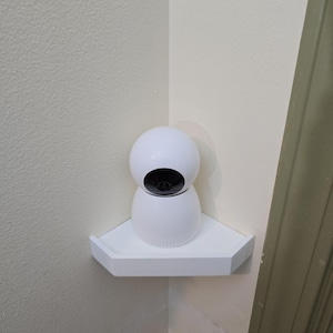 May include: A white security camera with a black lens sits on a white corner shelf. The camera has a rounded top and a cylindrical base. The shelf is mounted on a beige wall.