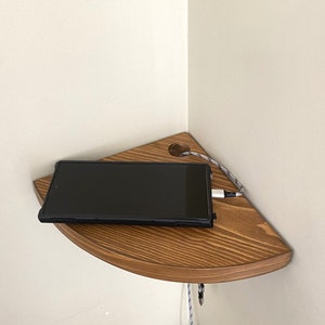 May include: A wooden corner shelf with a black phone on it, charging with a white cord. The shelf is mounted on a white wall.