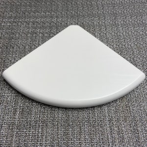 May include: White corner shelf made of a smooth, rounded material. The shelf is designed to fit in a corner and has a curved edge.