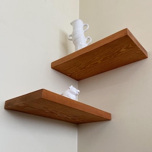 May include: Two wooden shelves mounted in a corner of a room. The top shelf has a white ceramic pitcher on it. The bottom shelf has a white ceramic figurine on it.