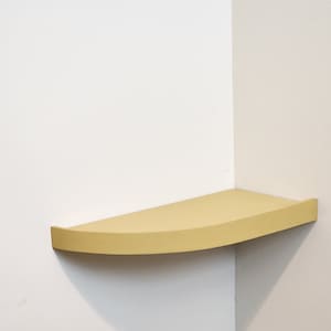 May include: A light yellow corner shelf mounted on a white wall. The shelf has a curved front edge and is designed to fit into a corner. The shelf is made of wood and painted a solid color.