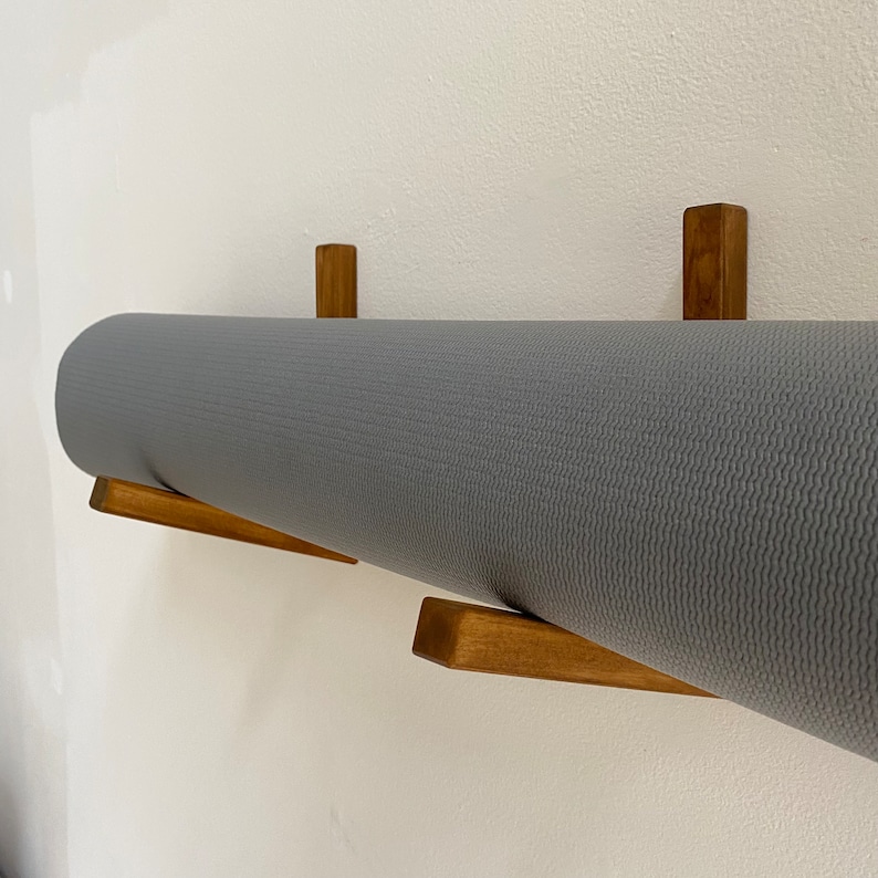 May include: A wooden yoga mat holder with two brown wooden brackets mounted on a white wall. A gray yoga mat is resting on the brackets.