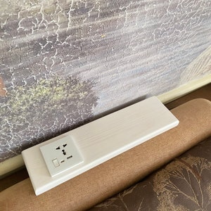 以下が含まれることがあります： 複数のプラグタイプとUSBポートを備えた白い電気コンセントが付いた白い木製の棚。 棚は長方形で、茶色の表面にあり、背景にはテクスチャ加工された絵画があります。