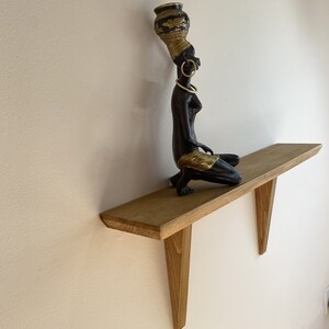 May include: A wooden shelf with a decorative figurine of a woman carrying a pot on her head. The shelf is mounted on a wall and has two triangular supports.