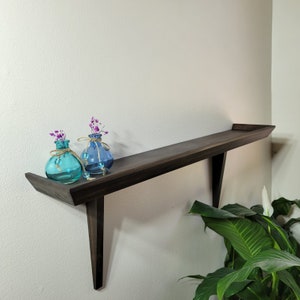 May include: A dark brown wooden floating shelf mounted on a white wall. Two small, round glass vases in shades of blue hold dried purple flowers. The shelf has a unique angled design.