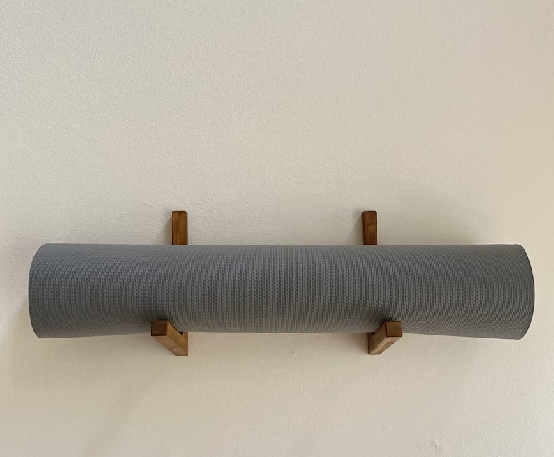 May include: A gray yoga mat is hanging on a wall mounted wooden rack. The rack is made of two pieces of wood that are connected at a right angle.