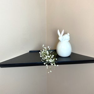 Puede incluir: Un estante de esquina negro con una figurita de conejo de cerámica blanca y una rama de flores blancas.