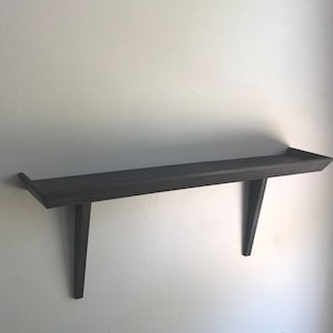 Könnte beinhalten: Ein dunkelgraues Holzregal an einer weißen Wand. Das Regal hat eine rechteckige Oberseite mit leicht erhöhtem Rand und wird von zwei abgewinkelten Halterungen getragen. Das Design ist minimalistisch und modern.