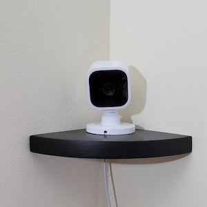 May include: A white security camera mounted on a black shelf. The camera has a round lens and a white base.