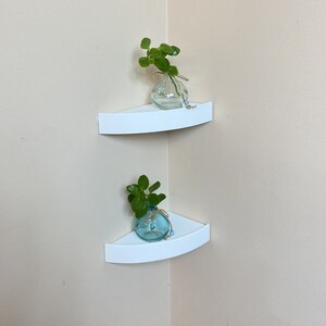 May include: Two white corner shelves with a curved front are mounted on a beige wall. Each shelf holds a small, clear glass vase with a sprig of green leaves. The vases are tied with twine and adorned with blue beads.