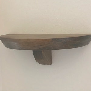 Puede incluir: Una estantería de madera marrón oscuro con un borde frontal curvo y un solo soporte. La estantería está montada en una pared blanca.