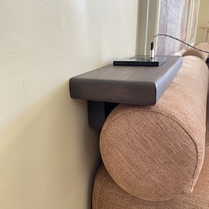 Puede incluir: Una estantería de madera marrón oscuro fijada a un sofá beige con una toma de corriente negra en la parte superior.