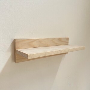 May include: A simple, light wood shelf mounted to a white wall. The shelf is made of two pieces of wood, one horizontal and one vertical, with a slight overhang on the front edge.