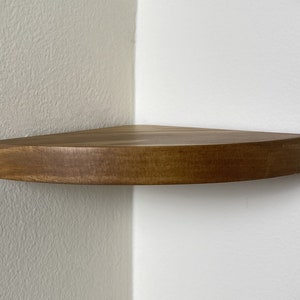 May include: A floating corner shelf made of stained wood. The shelf is a triangle shape with rounded edges and is mounted on a white wall. The wood has a warm, medium brown tone.