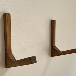 May include: Two wooden wall mounted L-shaped shelves. The shelves are made of dark brown wood and are mounted on a white wall.
